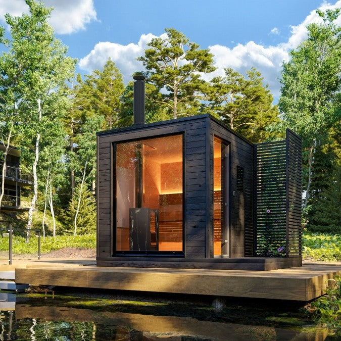 KUUT Wood Fired Sauna Premium on a raised lakeside deck, surrounded by forest, with glass front and warm sauna lighting.