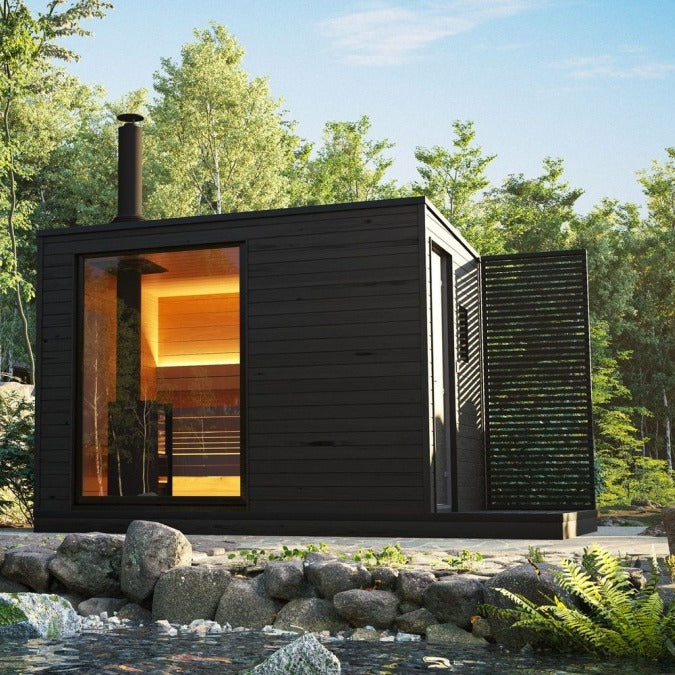 KUUT Wood Fired Sauna Premium unit beside a pond, featuring large front window and glowing interior lights, surrounded by trees.