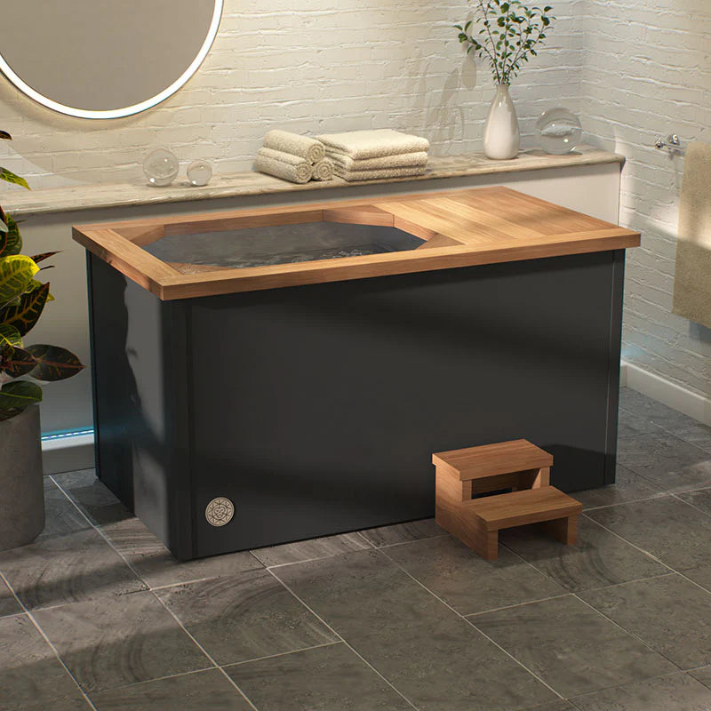 Brass Monkey Ice Bath with matte black body and wooden trim, accompanied by a wooden step stool, set in a bright indoor bathroom with tiled flooring and modern decor.