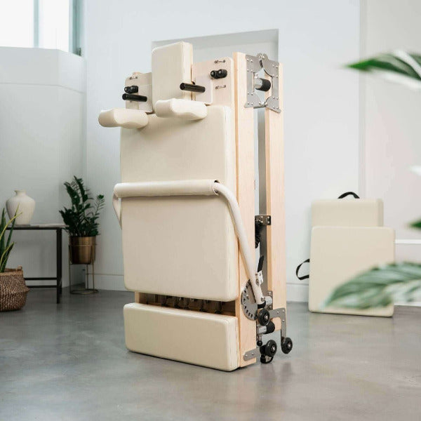 Beige foldable Pilates reformer upright against wall in modern studio.