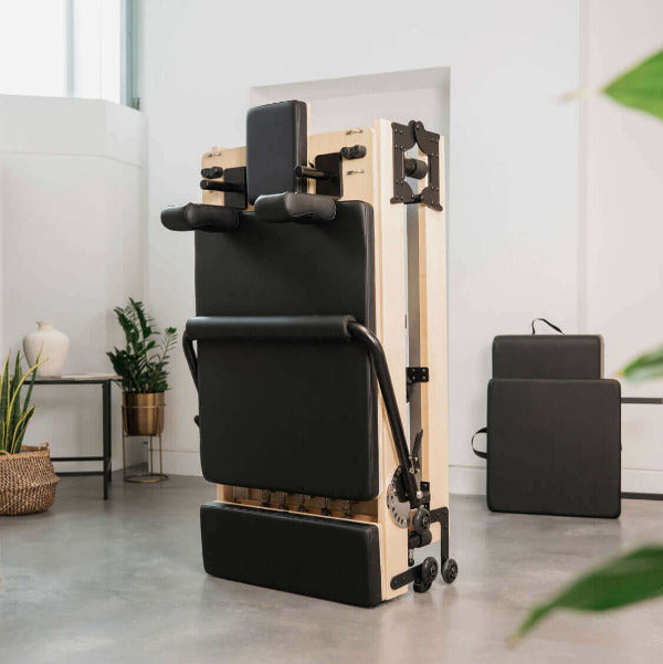 Black foldable Pilates reformer in upright storage position, maple frame.