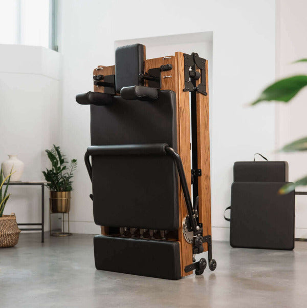 Black foldable Pilates reformer upright on red oak frame in modern studio.