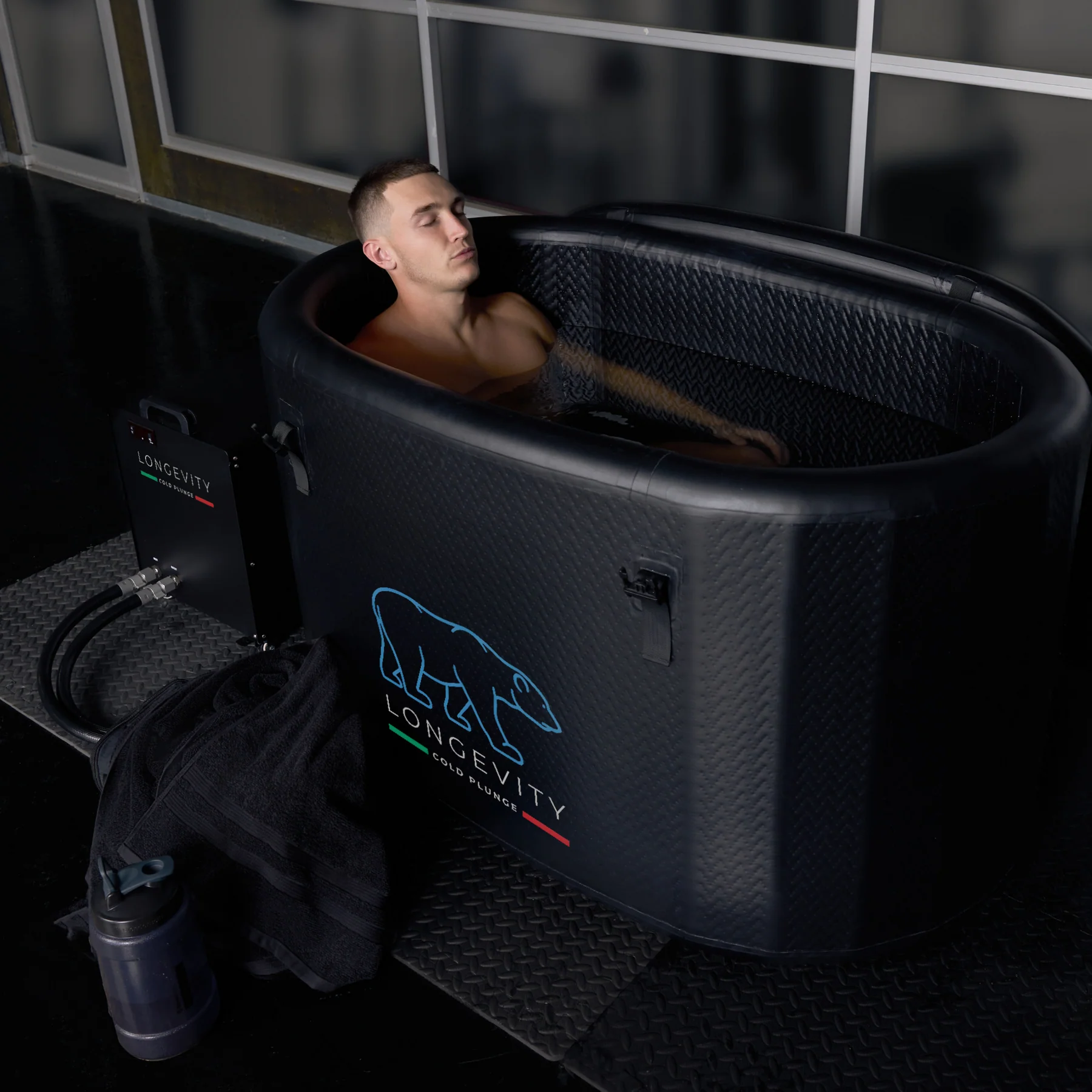 Man relaxing in filled Longevity Cold Plunge tub with towel and water bottle beside it.