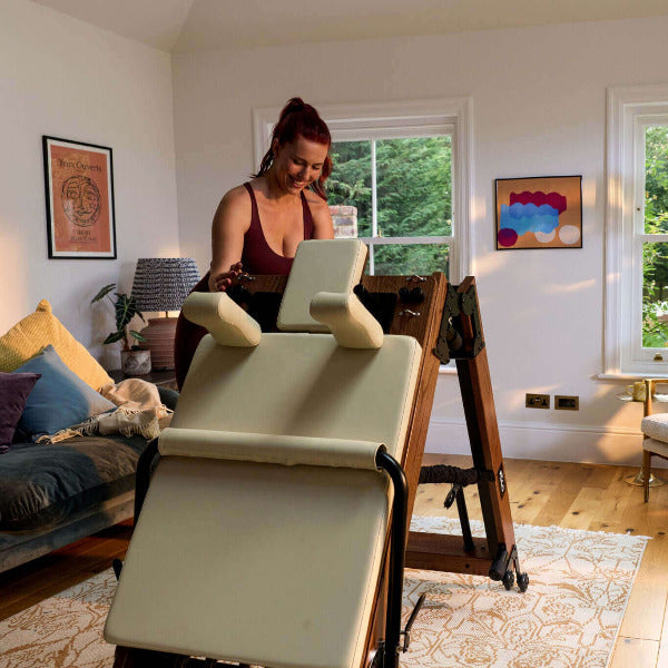 Beige foldable reformer with red oak frame being unfolded in bright living room.