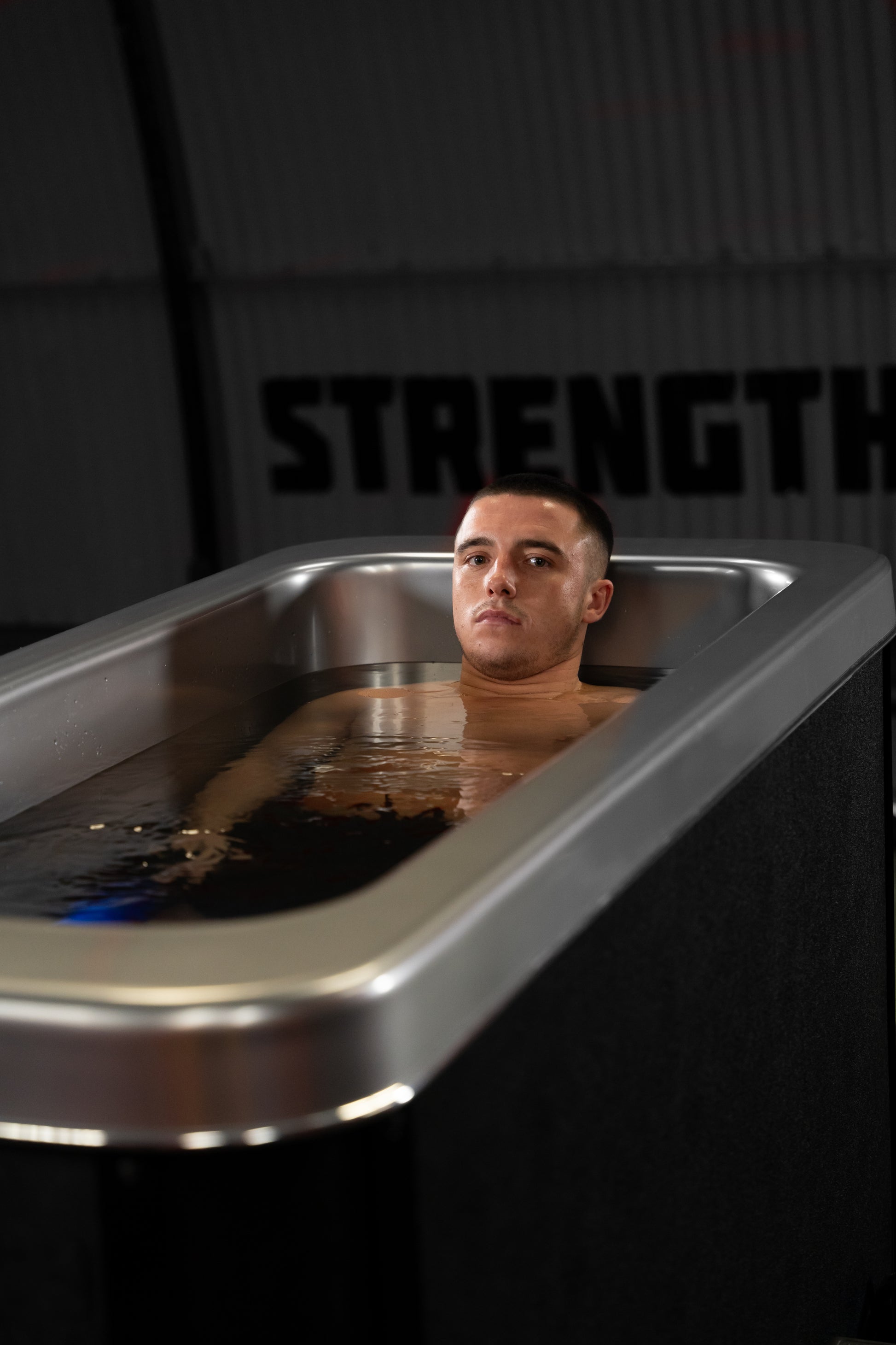 Man submerged in Optimal Health Ice Bath with focused expression, gym wall in background.