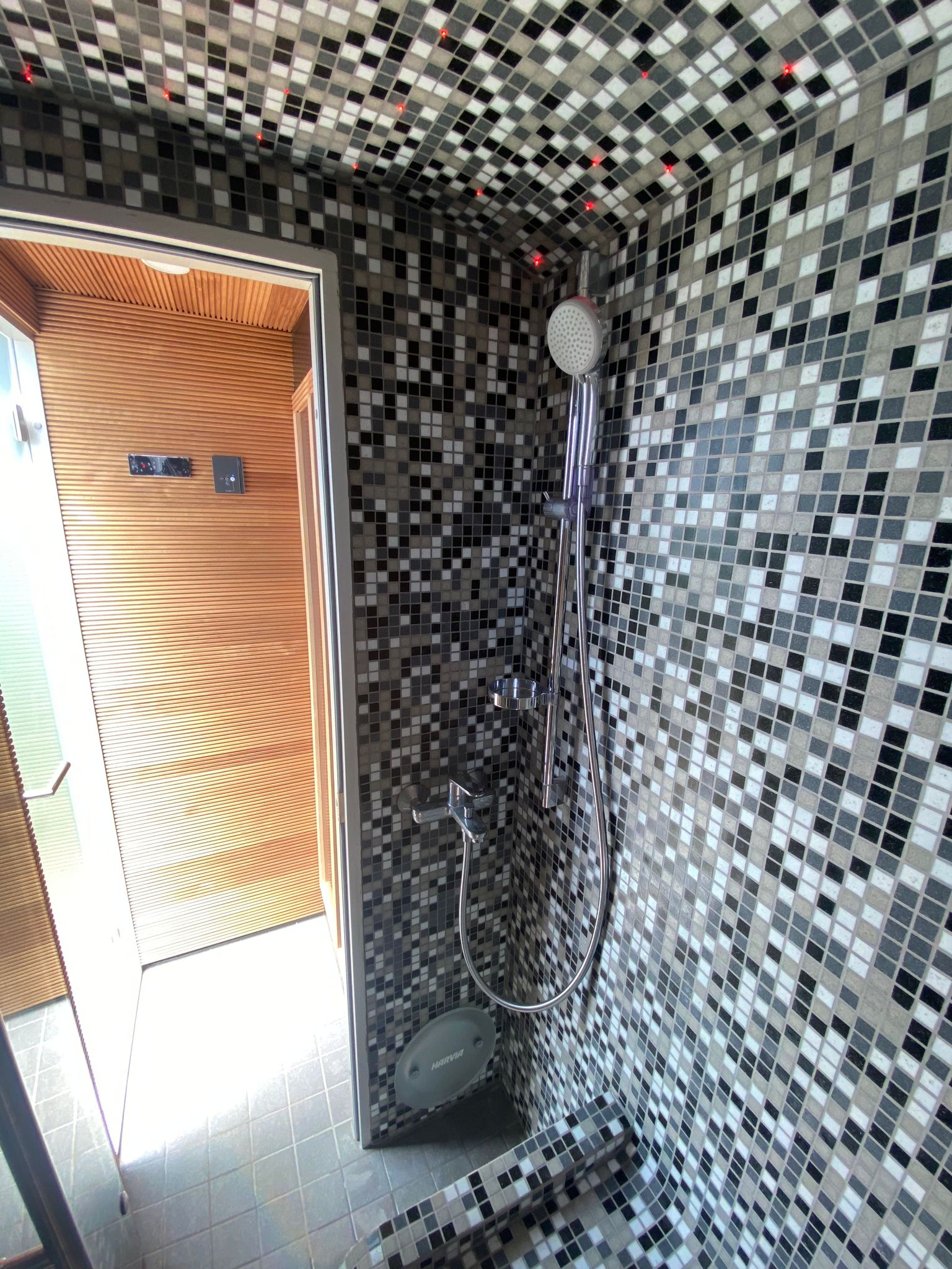 Interior of KUUT Outdoor Steam Sauna showing black and white mosaic tiled shower and wood-panelled steam room through glass door.