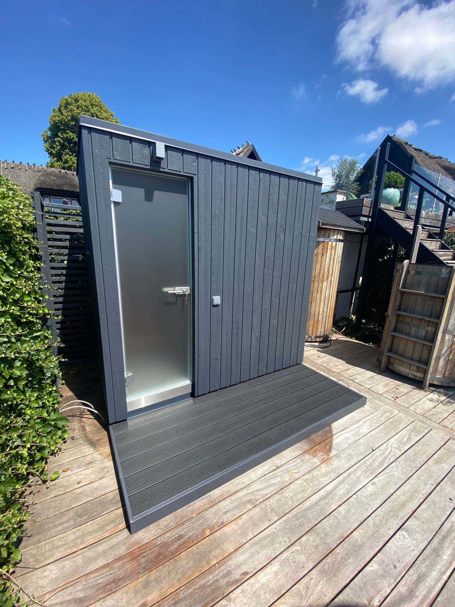 KUUT Outdoor Steam Sauna with dark wood cladding and frosted glass door, placed on a wooden deck under a clear blue sky.