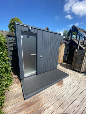 KUUT Outdoor Steam Sauna with dark wood cladding and frosted glass door, placed on a wooden deck under a clear blue sky.