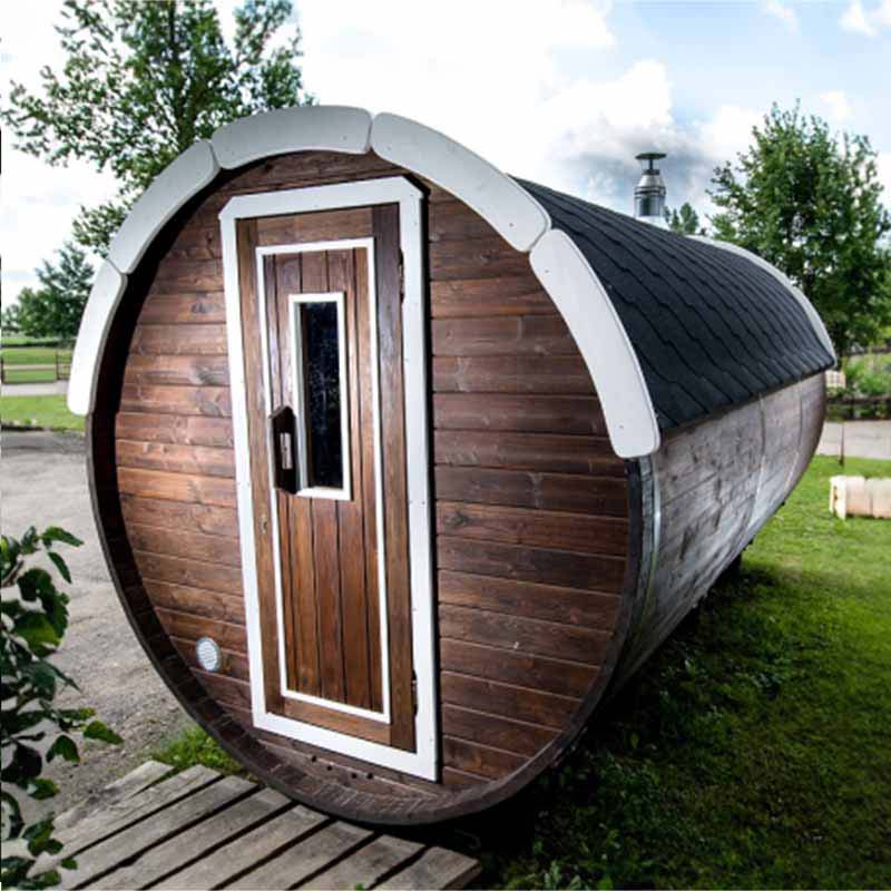 Front view of a dark wood barrel sauna with white-trimmed door outdoors.