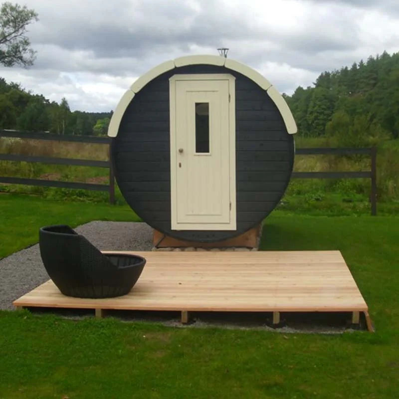 Outdoor black and white barrel sauna with wooden deck and chair on a grassy lawn.