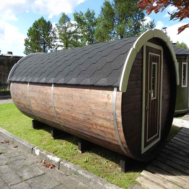 Side view of a dark wood barrel sauna with black shingle roof outdoors.